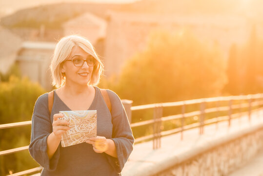 Adult female traveler standing on stone bridge and exploring map while checking direction for sightseeing during trip in Cuenca town in Spain in sunny day
