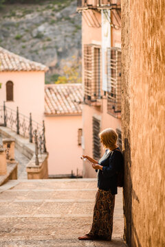 Positive side view of adult female tourist reading map and looking away with curiosity while standing against shabby stone wall of ancient building during sightseeing in old town Cuenca in Spain