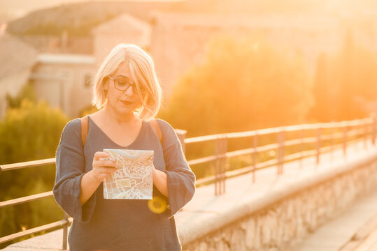 Adult female traveler standing on stone bridge and exploring map while checking direction for sightseeing during trip in Cuenca town in Spain in sunny day