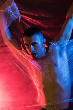 Aggressive male in casual clothing shouting and looking at camera while ripping transparent tape in light of red and blue lamps