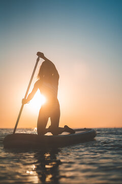 Unrecognizable silhouette of woman in swimsuit on SUP board in calm sea at sunset in summer