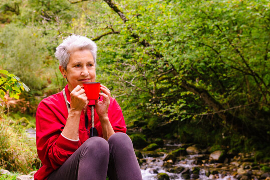Contemplative elderly female tourist drinking water near Casano river while resting on stone and looking away