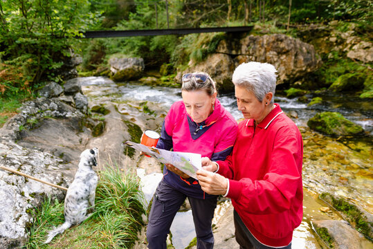 From above of middle aged female hikers watching paper map on mountain behind Casano river and dog in Spain