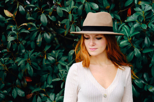 Attractive young long haired ginger female in stylish hat looking down while standing against green bushes in summer garden