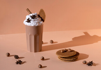 Glass with chocolate cocktail with whipped cream garnished with star anise and cinnamon stick placed on table with hazelnuts and cookies on pastel brown background