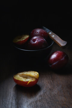 Still life composition with bowl with ripe aromatic juicy plums with water drops arranged on wooden table with knife and halved fruit