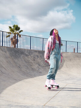 Delighted female in trendy street style outfit and with vivid dyed hair riding roller skates in skate park in summer while looking up