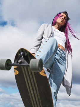 From below of confident female millennial with pink hair and in stylish clothes standing with longboard and looking at camera against blue sky