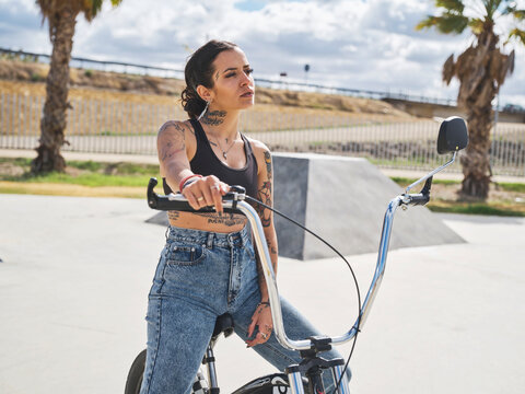 Determined tattooed female in stylish outfit sitting on BMX bicycle in skate park and looking away