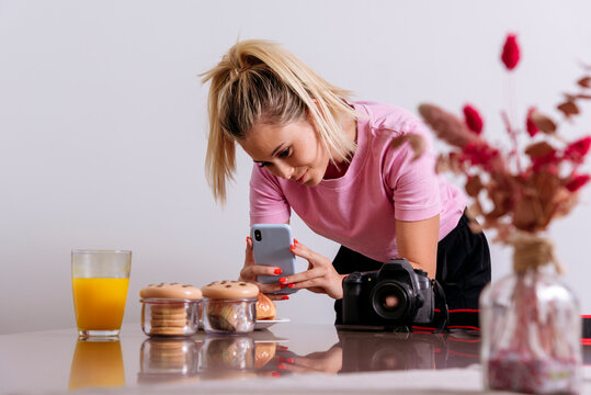 Female photographer taking picture of desserts and glass of orange juice with smartphone while working remotely from home