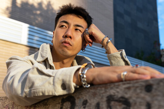 From below pensive Asian male in casual wear sitting near street stone fence and leaning head on hand while looking away thoughtfully