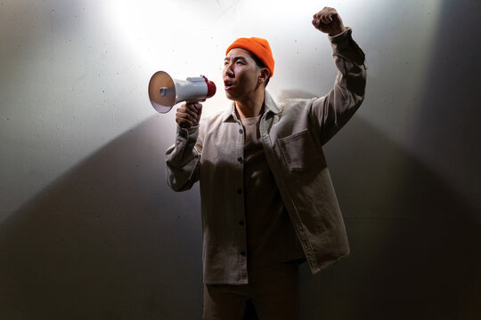 Side view of overwhelmed male protesting in casual clothes screaming in loudspeaker while standing against gray wall in dark room