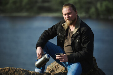 Portrait of an adventurer man sitting on rocks and holding thermos with hot drink and having a rest while hiking in nature.