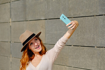 Trendy young red haired female in stylish hat standing near stone building and taking selfie on mobile phone