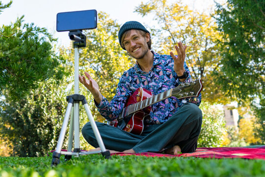 Ground level of handsome man sitting with electric guitar in park and recording video on smartphone for blog on social media