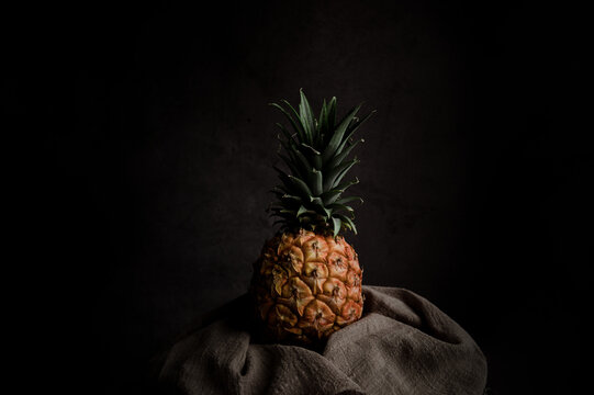 Still life composition with whole ripe pineapple wrapped in textile on wooden stand against black background