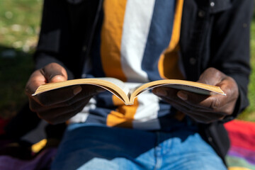 Cropped unrecognizable ethnic male sitting on blanket and reading interesting novel while relaxing on summer day in park