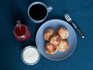 Pancakes made from cottage cheese on a gray plate with sour cream and strawberry sauce and coffee with fork on a table. Tasty breakfast concept
