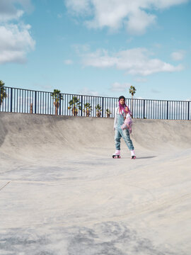 Delighted female in trendy street style outfit and with vivid dyed hair riding roller skates in skate park in summer while looking at camera