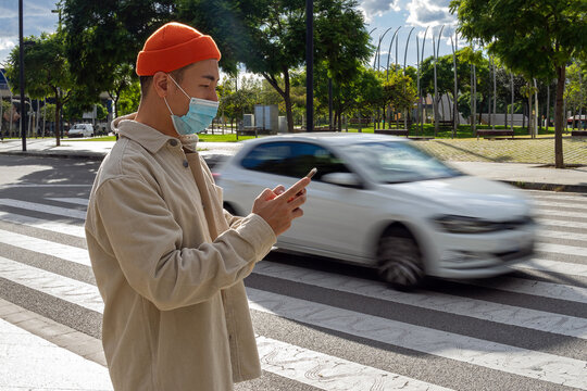 Side view focused young Asian male wearing casual outfit and orange hat browsing mobile phone while standing on sunny street near moving car