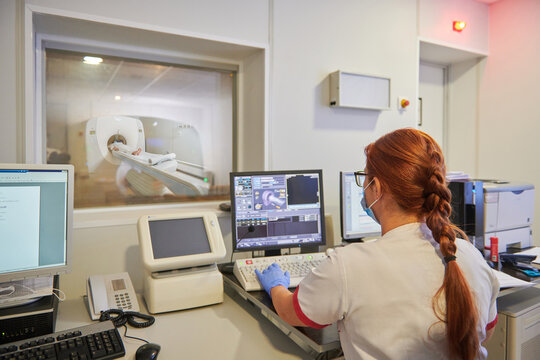 Back view of anonymous female doctor in uniform using desktop computer while realizing tomography test with patient on equipment