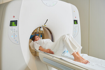 Barefoot patient in white wear lying with closed eyes on couch of tomography machine in hospital