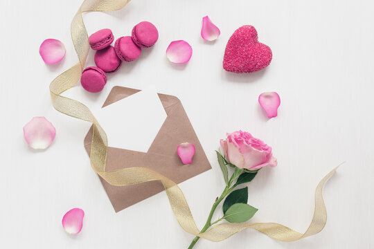 Blank greeting card mockup in envelope, rose, macaroon, gold ribbon, heart and rose petals. White painted wood background. Valentine's Day. Template for your design. Top view, flat lay.