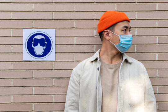 Serious Asian male wearing mask standing near sign building wall illustrating person in protective mask looking away