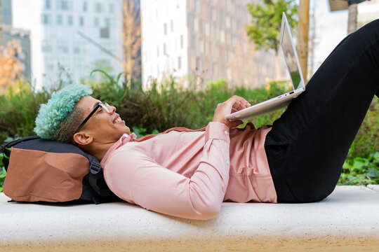 Side View Of Smiling African American Male Student With Blue Hair Lying On Backpack In University Campus And Typing On Laptop While Working On Homework Assignment