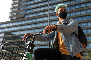 Low angle of black male hipster in protective mask riding modern bike in city during coronavirus outbreak and looking away
