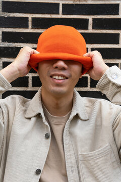 Happy male putting knitted orange hat on head and covering eyes while standing against black brick wall