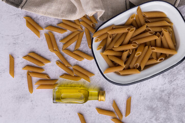 From above of dried Penne pasta in metal bowl and bottle of virgin olive oil arranged on table with napkin in kitchen