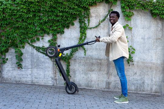 Side view of positive African American male standing on sloping street with contemporary electric scooter and looking at camera while having fun
