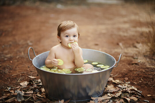 Cute Baby Enjoying Fresh Lemon Slices While Sitting In Metal Bath With Water On Sandy Land And Looking At Camera