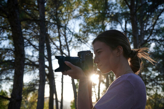 Side view of young female photographer with professional photo camera shooting pictures while standing in green forest in sunny day