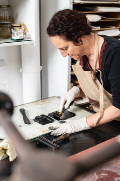 Female artisan in glove using braver and spreading ink on sheet for engraving while working in professional workshop