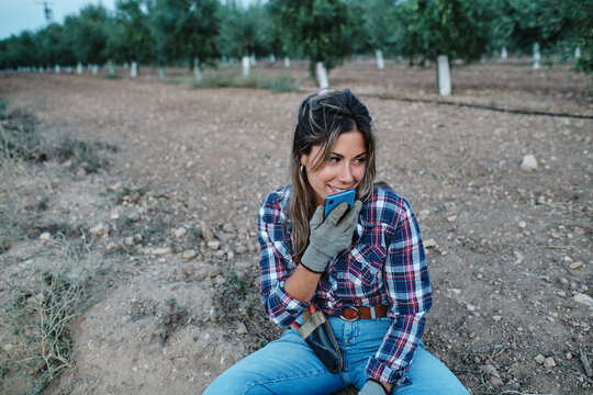 Female farmer sitting on ground and recording audio message via mobile phone while relaxing during work and looking away