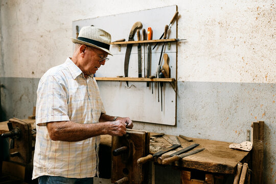 Side view of senior male carpenter grinding wooden details while making handmade folding fans in messy workshop