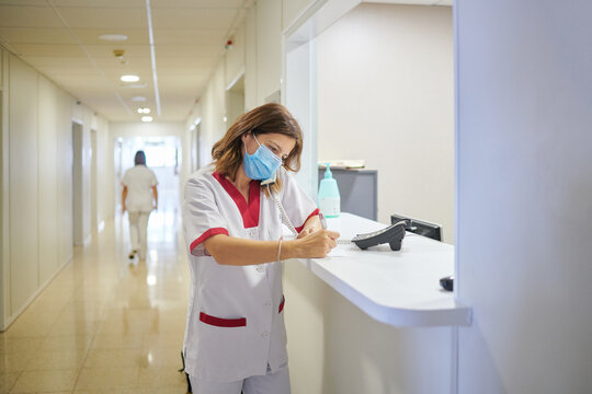 Concentrated female nurse wearing white medical suit and face mask taking notes in clinical record chart while standing near modern hospital reception