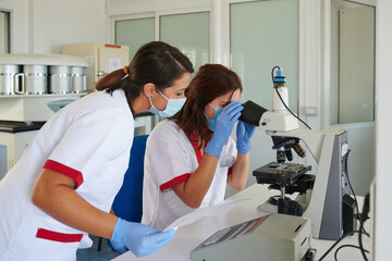 Unrecognizable female medical technologists in uniforms and sterile masks working with microscope in laboratory