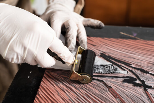Closeup of crop anonymous master in gloves with professional roller applying black ink on engraved plate during printmaking process in workshop
