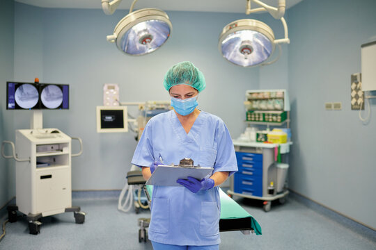 Anonymous female coworker in medical uniform and mask interacting while revising papers and standing behind shiny lamps in clinic