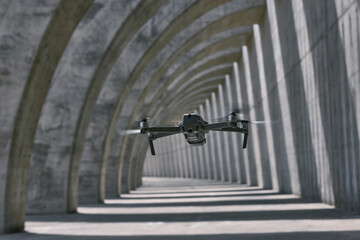 Modern drone flying in empty arched walkway with columns casting shadows in shape of lines on sunny day