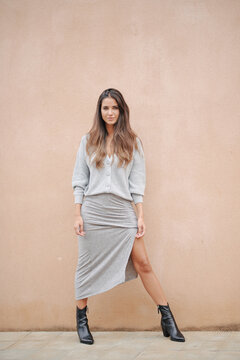 Charming female with long hair and in casual dress standing near building on street and modestly looking at camera