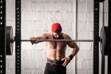 Strong male athlete with naked torso and in weightlifting belt standing near barbell during workout in gym