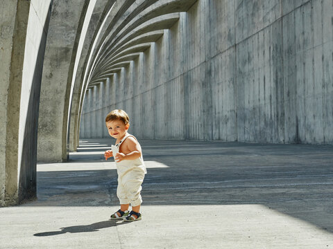 Delighted little kid in cute overalls walking along arched pathway in city and looking at camera
