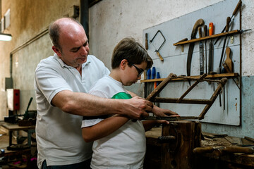 Side view of father teaching son using metal rasp for grinding wood in grungy dirty woodwork workshop