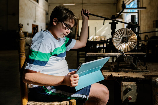 Side view of teen boy sitting at workbench in shabby workshop and using mechanism for producing folding fans while watching video on tablet