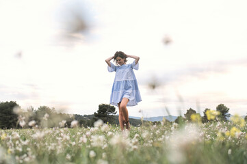 From below full body delighted female in short blue dress walking through meadow covered with grass and flowers next to forest and mountain valley under cloudless sky in daytime
