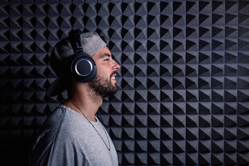 Side view of stylish male singer wearing headphones standing in recording studio on background of soundproof acoustic panel and looking at camera
